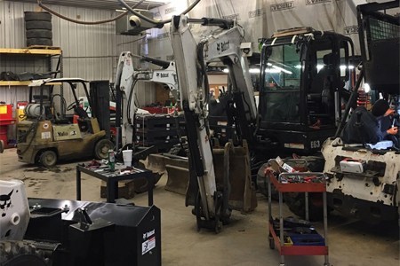 Two white Bobcat® excavators inside a garage with repair supplies and a dark green forklift nearby.