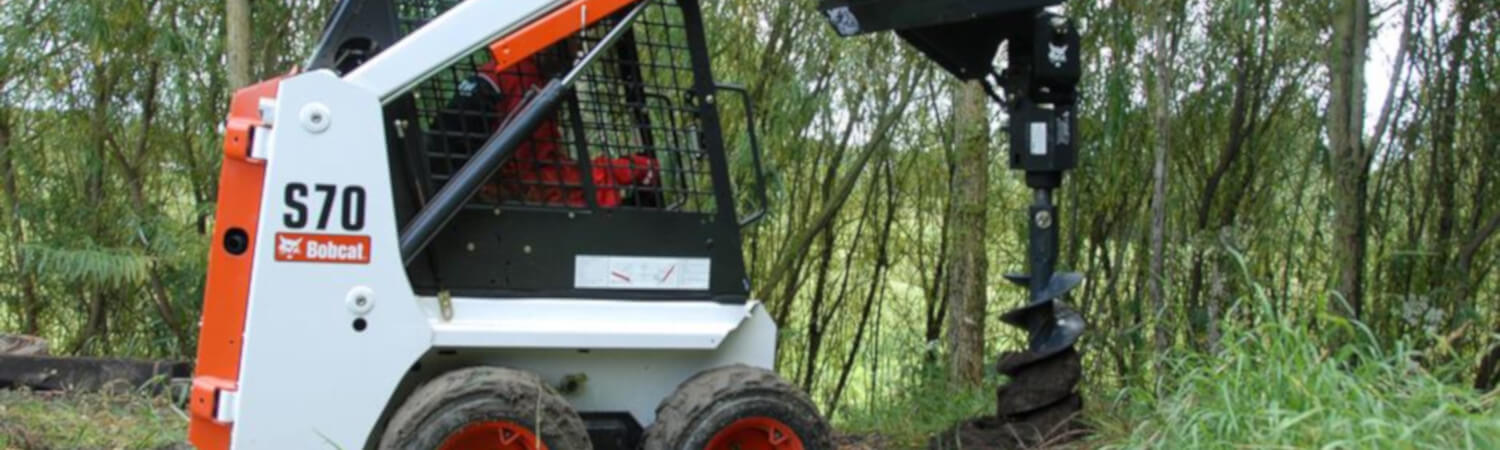 Bobcat® S70 skid steer with an auger attachment drilling into the ground in a dense forest.